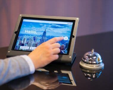 Ruka dodiruje ekran tableta za prijavu gostiju na hotelskoj recepciji, pored njega zvonce